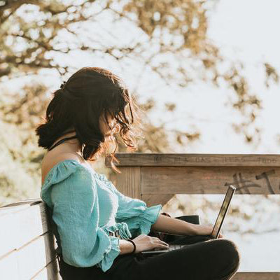 yound woman using her computer while in a wooded area. she's sitting on a wooden bench.