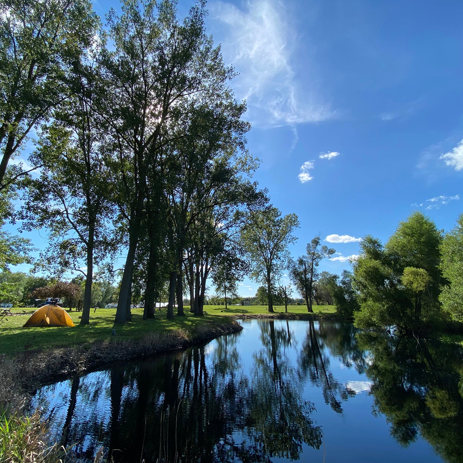 view over pond with sky reflecting in water. tent in background on a site next to water.